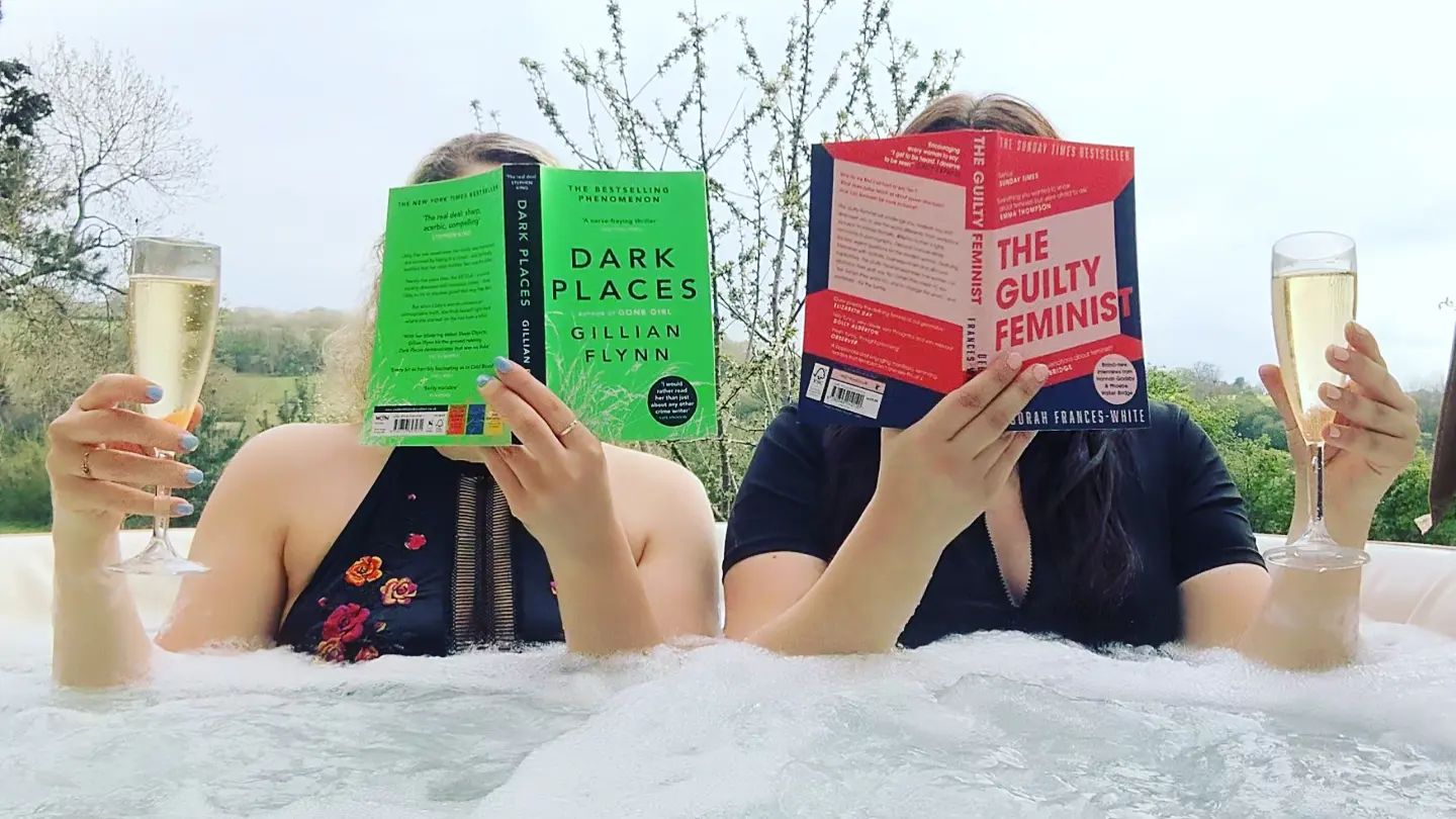 Two women reading books in an outdoor hot tub while holding glasses of sparkling win